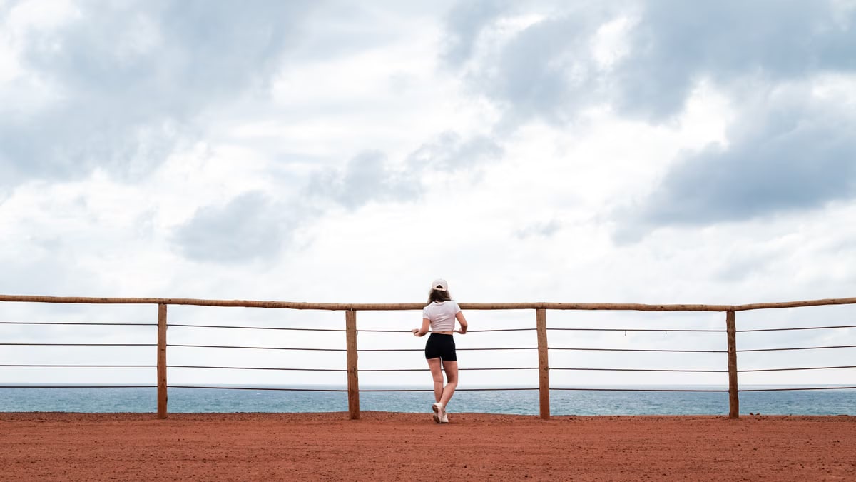 A woman stands on a red sand cliff overlooking the ocean, under a cloudy sky in Natal, Brazil.